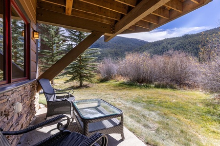 Outdoor patio with two wicker chairs and a glass table, overlooks a grassy area and mountainous landscape with trees under a clear blue sky.