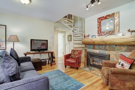 A cozy living room with a stone fireplace, blue sofa, two armchairs, and a television on a wooden table. A spiral staircase leads to an upper floor. The room is warmly illuminated.