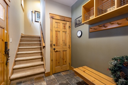 An entryway features a wooden bench, coat hooks, a clock, and a welcome sign. A staircase leads upward, and a wooden door is in the center. A small wreath is visible on the right.