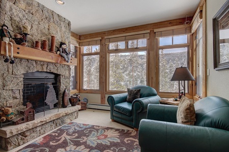 A cozy living room with large windows showing a snowy forest view. It features a stone fireplace with decorations, two green armchairs, a lamp, and a colorful patterned rug.