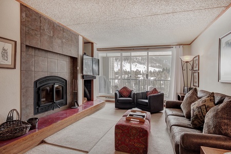 A cozy living room features a fireplace, a mounted TV, armchairs, a couch, and a large window with a snowy mountain view. The room is decorated with framed art and a floor lamp.