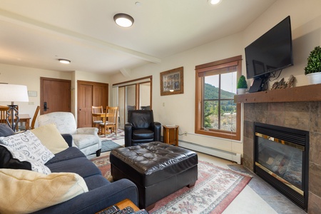 A cozy living room with a fireplace, wall-mounted TV, large window with a scenic view, black leather ottoman, beige and black sofas, and a dining area in the background.