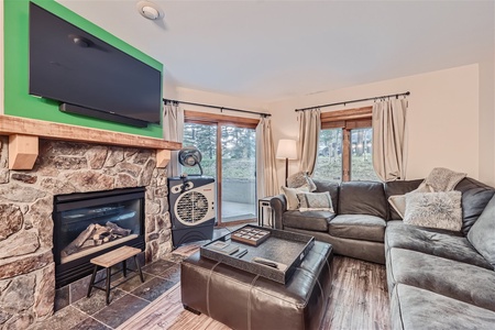 A cozy living room with a stone fireplace, mounted TV, corner sofa, small table, ottoman, window with curtains, and a floor lamp. The room has large windows allowing natural light in.