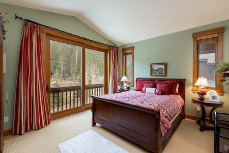 A bedroom with a large wooden bed, red and white bedding, two nightstands with lamps, large windows, and green walls. The room opens to a balcony with a forest view.