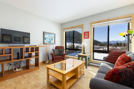 A living room with a sofa, armchair, wooden coffee table, TV on a wooden stand, and large windows showcasing a scenic mountain view.