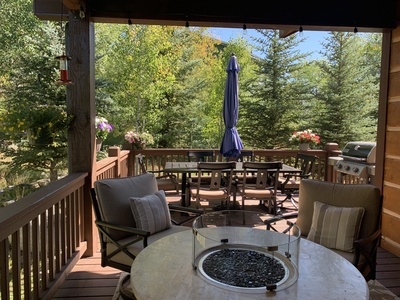 A wooden deck features outdoor furniture, a gas fire pit, a dining table with an umbrella, a grill, and potted flowers surrounded by trees and greenery.