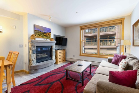 A cozy living room with a stone fireplace, a large window, a beige couch with red cushions, a wooden coffee table, and a flat-screen TV. The room features wooden furniture and a red patterned rug.