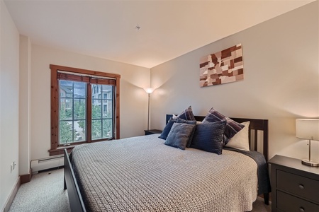 A neatly made bed with multiple pillows and a textured blanket is in a minimalist bedroom with two bedside tables, a standing lamp, and a window. An abstract artwork is hung above the bed.