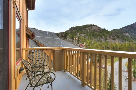 A balcony with three metal chairs overlooks a scenic view of forested hills and distant houses under a cloudy sky.