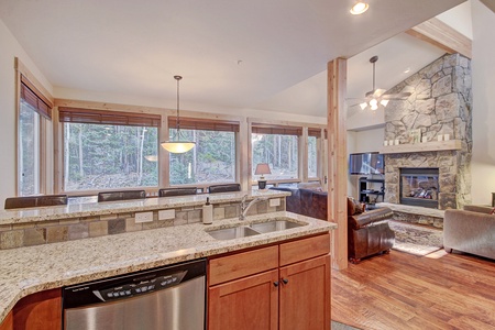 Spacious, modern kitchen and living area with granite countertops, wooden cabinetry, large windows, forest view, stone fireplace, and hardwood floors.