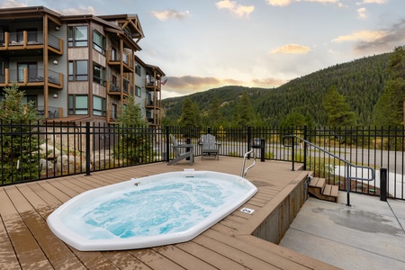 An outdoor hot tub is situated on a wooden deck by a modern apartment complex with forested hills and a cloudy sky in the background.