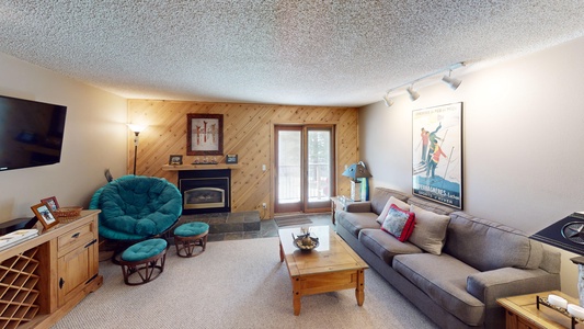 Cozy living room with a TV, fireplace, cushioned chairs, a wooden coffee table, and a sofa. The walls are adorned with pictures and posters, with glass doors leading to an outdoor area.