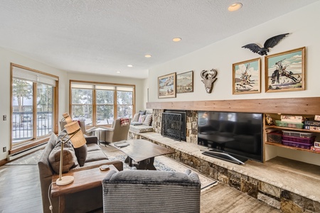 A cozy living room with large windows, stone fireplace, mounted TV, and various decorations including wall art, a stuffed bird, and shelves with board games.