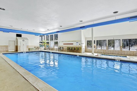 Indoor swimming pool area with blue water, lounge chairs, and large windows providing natural light. The space features tiled walls, a high ceiling, and safety equipment along the pool's edges.