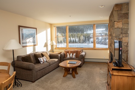 A cozy living room with a brown sofa, wooden coffee table, TV on a stand, and large window with a scenic outdoor view. The room is decorated with lamps, a wall painting, and has a stone accent wall.
