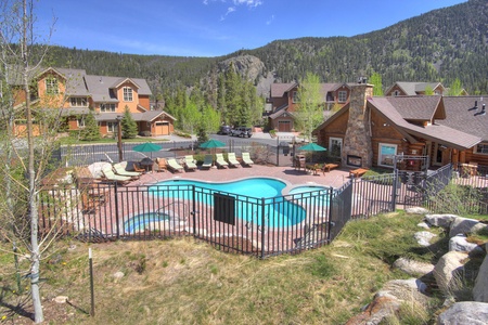 A mountainside resort with a fenced outdoor pool area, lounge chairs, umbrellas, and several rustic-style buildings surrounded by trees and hills.