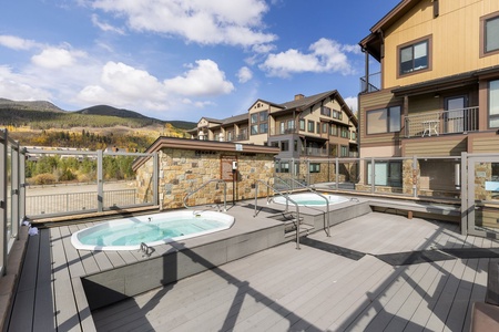 An outdoor area with two raised hot tubs, surrounded by a wooden deck and glass railing. There are multi-story buildings and mountains in the background under a partly cloudy sky.