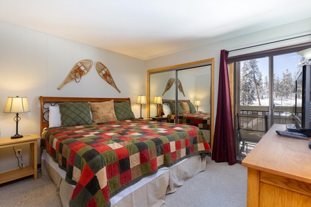 A bedroom with a quilted king-size bed, two bedside tables with lamps, a mirrored closet, a dresser with a TV, and a window with a view of a snowy landscape. Snowshoes are mounted above the bed.