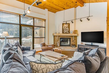 A cozy living room with a large sectional sofa, a wooden coffee table, a fireplace, and a flat-screen TV. The room features a high wooden ceiling and large windows allowing natural light to enter.