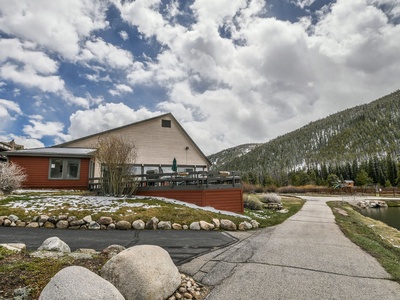 A house with a wraparound deck is situated near a narrow road, surrounded by rocks and partial snow coverage. A forested mountain is visible in the background under a cloudy sky.