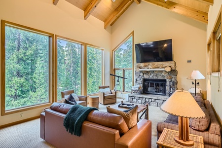 A cozy living room with large windows offering forest views, featuring a stone fireplace, a flat-screen TV, a leather sofa, a wicker chair, and wooden ceiling beams.