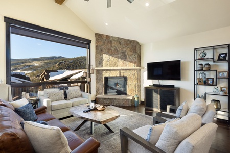 A cozy living room with a large window, stone fireplace, television, and comfortable seating. The room has a rustic decor and offers a scenic mountain view through the window.