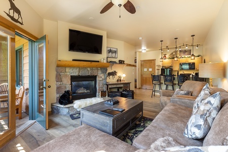 Living room with a stone fireplace, mounted TV, and sectional sofa. Wooden coffee table in center. Open concept connects to the kitchen with bar stools. Balcony door open, showing a patio.