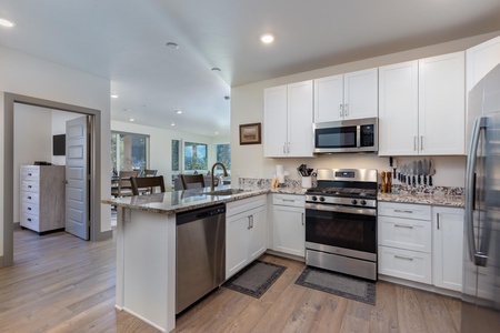 Modern kitchen with white cabinets, stainless steel appliances, granite countertops, and wood flooring. Room includes a dining area and is adjacent to a living space visible through a doorway.