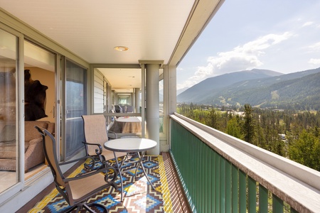 A balcony with two chairs and a table overlooks a scenic view of mountains and trees. The balcony has a patterned rug and sliding glass doors leading to an indoor area.