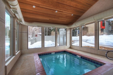 Indoor heated pool with tile surround, brick trim, and large windows offering a view of a snow-covered outdoor area. Wooden ceiling with recessed lights.