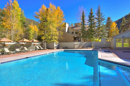 A clear blue outdoor swimming pool is surrounded by lounge chairs and beige umbrellas, set against a backdrop of autumn trees and a building with a mountain behind it.