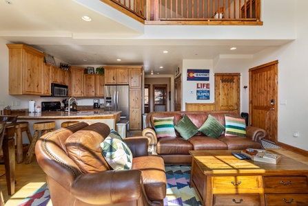 Living room with leather sofas, wooden coffee table, and open kitchen. Wooden cabinets and decor create a rustic feel. A poster on the wall depicts skiing imagery.