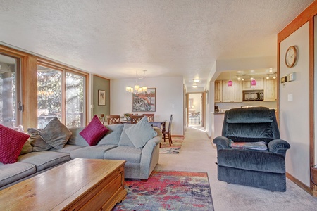 Living room with gray sectional sofa, colorful cushions, wooden coffee table, and reclining chair. Dining area and kitchen visible in the background. Large windows let in natural light.