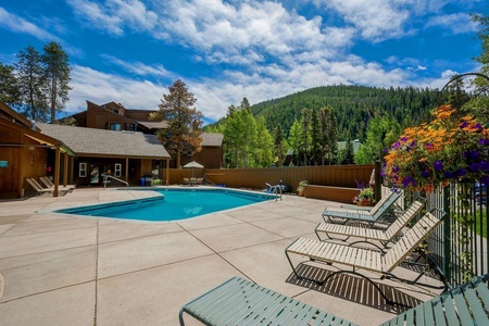 Outdoor pool area surrounded by lounge chairs and wooden buildings with a forested mountain in the background under a blue sky with some clouds.