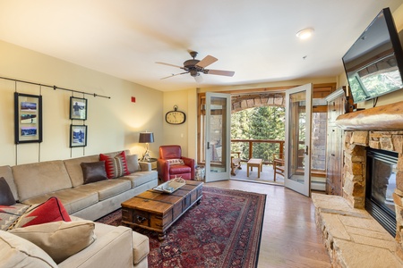 A living room with a red armchair, beige sofa, wooden coffee table, wall-mounted TV, and fireplace. Open double doors lead to a balcony with outdoor seating and view of trees.