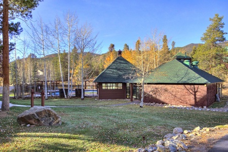 A brown wooden cabin with a green roof is situated among trees and a lawn, near a fenced pool, with mountains visible in the background under a clear blue sky.