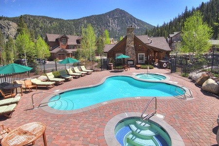 Outdoor pool area in a mountain resort, featuring a kidney-shaped pool, a hot tub, lounge chairs, green umbrellas, and a wooden lodge. Trees and mountains are visible in the background.