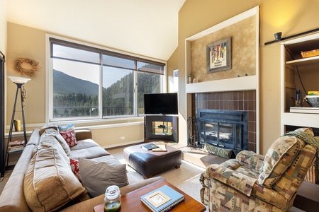 A cozy living room with a large window showcasing a mountain view, a TV beside a fireplace, a beige sofa, patterned armchair, and a glass jar on a small table. Bookshelves are built into the wall.