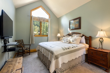 A cozy bedroom with a sloped ceiling features a double bed, two bedside tables with lamps, a wall-mounted TV, a wooden chair, a window with a view of trees, and a rustic wooden chest.