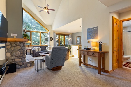 A cozy living room with vaulted ceilings, stone fireplace, grey armchair, wooden coffee table, and large windows. A dining area with a chandelier is visible in the background and a bathroom is on the right.