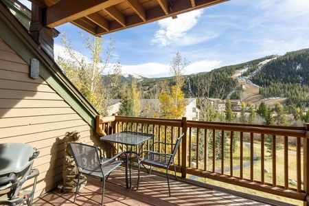 A wooden balcony with a glass table and metal chairs, offering a view of green and yellow trees and a mountainous landscape under a partly cloudy sky. A grill is also visible on the left side.