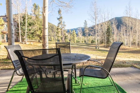 Outdoor patio with a round metal table and four mesh chairs, set on a green mat. The setting overlooks a scenic landscape of sparse trees, grassy fields, and distant mountains.