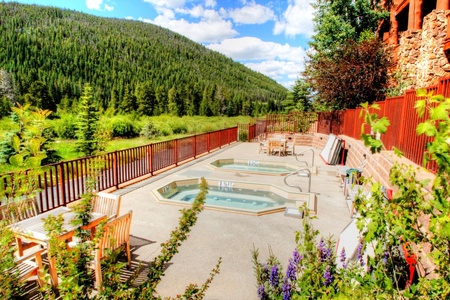 Outdoor hot tub area with wooden seating, surrounded by lush greenery and mountains in the background under a partly cloudy sky.