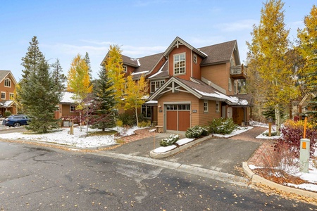 Two-story brown house with white trim surrounded by trees with autumn foliage and a thin layer of snow on the ground, situated at a street corner.