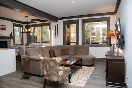 A cozy living room featuring a sectional sofa, an armchair, a coffee table, a wall-mounted TV, and large windows with views of trees.