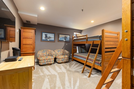 A basement bedroom features a bunk bed, two cushioned chairs, wooden furniture including a chest and a desk, framed artwork on the walls, and a flat-screen TV.