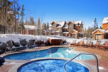 A view of an outdoor swimming pool and hot tub surrounded by snow with lounge chairs on the pool deck and houses in the background under a clear blue sky.