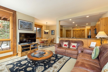 A cozy living room with a leather sofa, TV on a wooden stand, and a small coffee table. The space opens into a dining area with a wooden table and chairs. A kitchen with wooden cabinets is visible.