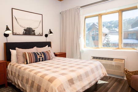 A cozy bedroom features a bed with plaid bedding, two red nightstands, and a wall-mounted art piece above the headboard. A large window reveals a snowy outdoor scene.