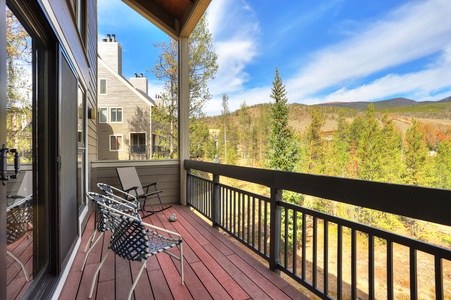 A wooden balcony with two woven chairs and a lounge chair overlooks a scenic mountain landscape with trees under a blue sky and white clouds.